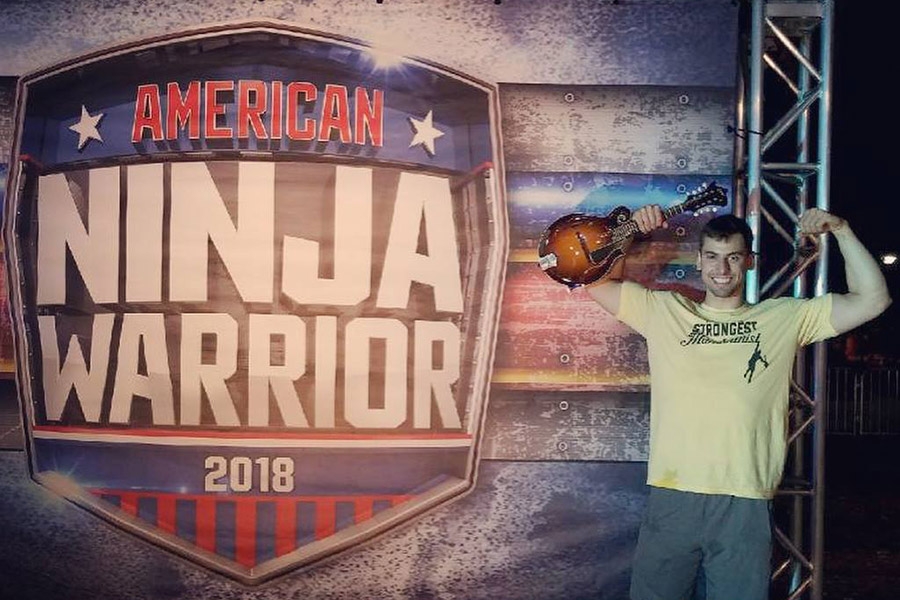 Civil engineering undergrad Elie Cohen flexes his biceps while holding his mandolin in front of an American Ninja Warrior 2018 sign. (Photo Courtesy: NBC)
