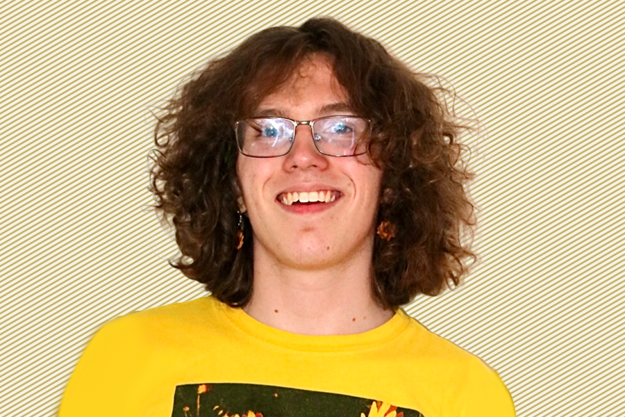 A photo of Hudson McGaughey wearing a yellow T-shirt and glasses in front of a gold and white striped background